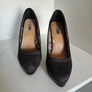 Svarta pumps från Din Sko, mocka-look - Snygga svarta pumps från Din Sko med mockaliknande yta och höga, blanka klackar. Skorna har spetsig tå och en plattform framtill. Insidan är fodrad med ett färgglatt blommigt mönster som ger en cool kontrast till den svarta utsidan.
