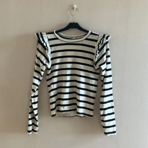 Randig långärmad tröja med volang - Snygg långärmad tröja med svartvita ränder och volangdetaljer på axlarna. Tröjan har en klassisk rund halsringning och är figurnära i modellen. Perfekt för dig som gillar en trendig och lekfull stil. Säljes pga av urväxt 