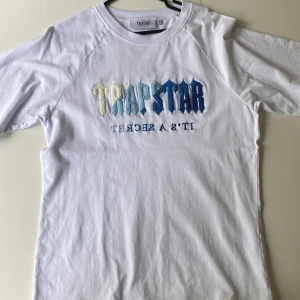 Trapstar T-Shirt - Vit t-shirt från Trapstar i storlek L med broderad logga i olika blå och vita nyanser på bröstet. T-shirten har korta ärmar och rund hals. Materialet är mjuk bomull och passformen är normal. Snyggt broderad text 'IT'S A SECRET' under loggan.