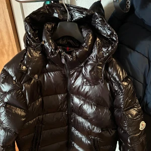 Svart Moncler pufferjacka med huva - Säljer en svart, glansig pufferjacka från Moncler med huva och dragkedja. Jackan har klassisk quiltad design, två fickor med dragkedja och Moncler-logga på ärmen. Perfekt för kalla dagar och riktigt snygg streetstil.                                                   Priset kan diskuteras 