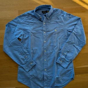 Blå skjorta från Polo Ralph Lauren - Klassisk ljusblå skjorta från Polo Ralph Lauren med button-down krage och broderad gul logga på bröstet. Skjortan har lång ärm, knappar framtill och är tillverkad i mjuk bomull. Perfekt för en clean och stilren look.