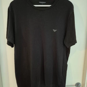 Svart t-shirt från Emporio Armani XL - Säljer en svart t-shirt från Emporio Armani med diskret logga på bröstet. Klassisk rund halsringning och korta ärmar. Tillverkad i mjuk bomull som känns skön mot huden. Perfekt för dig som gillar stilrena och enkla plagg med exklusiv känsla.
