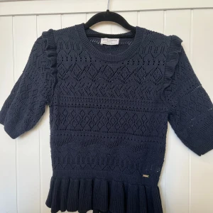 Mörkblå stickad topp Holly & Whyte - Supersöt mörkblå stickad topp från Holly & Whyte med kort ärm, volangdetaljer på axlarna och bred volangkant nertill. Snyggt hålmönster över hela tröjan och rund halsringning. Perfekt för dig som gillar unika detaljer och en feminin vibe.