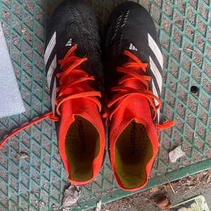 Adidas Predator fotbollsskor svart/röd - Säljer ett par Adidas Predator fotbollsskor i svart med röda detaljer och vita ränder. Skorna har snörning, mönstrad ovandel och fasta dobbar för gräsplan. Insidan är gul och modellen är Predator 20. Perfekta för dig som vill sticka ut på planen.