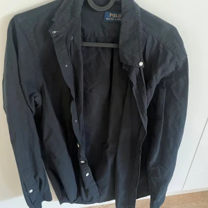 Svart skjorta från Polo Ralph Lauren - Svart långärmad skjorta från Polo Ralph Lauren med klassisk krage och knappar framtill. Skjortan har diskret broderad logga på ärmen och är tillverkad i bomull. Perfekt för en clean och stilren look.