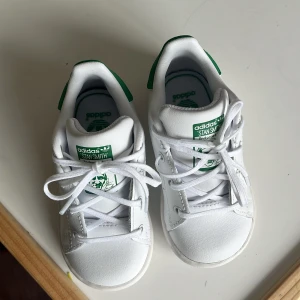 Adidas Stan Smith vita sneakers barn - Klassiska Adidas Stan Smith sneakers i vitt skinn med gröna detaljer på häl och plös. Snygg och tidlös modell med snörning och rund tå. Perfekta för en cool och sportig look. Mjuk innersula med Ortholite för extra komfort.