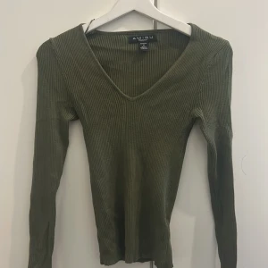 Olivgrön ribbad tröja från Amisu - Snygg olivgrön ribbad tröja från Amisu med v-ringad hals och lång ärm. Tröjan är figurnära och har en mjuk, stickad känsla som passar perfekt till jeans eller kjol. Enkel men trendig design som funkar året runt.