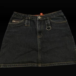 Mörk blå jeanskjol med dragkedjor - Cool marinblå jeanskjol med flera dragkedjor, D-ringar och bälteshällor. Kjolen har detaljer i silver och kontrastsömmar i gult. Kort modell med rå kant nertill och extra zip-detalj i sidan för edgy vibe.