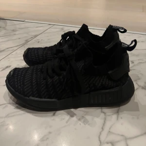 Svarta Adidas NMD sneakers - Snygga svarta Adidas NMD sneakers med stickad ovandel och klassiska detaljer. Skorna har svart sula, snörning och dragflikar med text bak. Perfekta för dig som gillar streetstyle och bekväma skor med modern design.