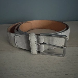 Beige mockabälte med silverfärgat spänne - Stilrent beige bälte i mocka med ett klassiskt silverfärgat metallspänne. Bältet har en mjuk yta och enkel design som passar till jeans eller chinos. Perfekt accessoar för att lyfta din outfit med en touch av lyxig känsla.