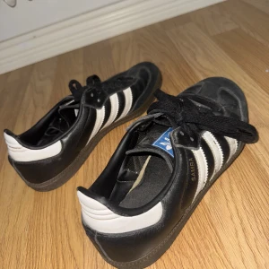 Adidas Samba svarta sneakers - Klassiska Adidas Samba sneakers i svart skinn med vita detaljer och de ikoniska tre ränderna på sidorna. Skorna har svart snörning, platt sula och rund tå. 