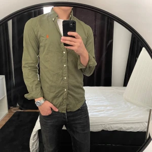 skjorta från Ralph Lauren - Säljer en olivgrön skjorta från Ralph Lauren i slim fit med klassisk krage och knappar hela vägen. Skjortan är i tunn bomullstwill och har den ikoniska broderade loggan på bröstet. Perfekt för dig som gillar stilrena och snygga plagg.
