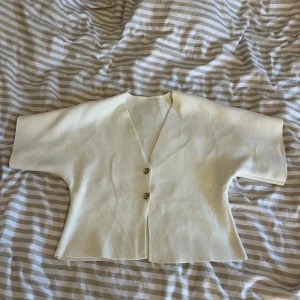 Vit  kofta med kort ärm - Superfin vit kofta med korta ärmar och v-ringning. Koftan har två snygga guldfärgade knappar framtill och är ribbad i materialet. 