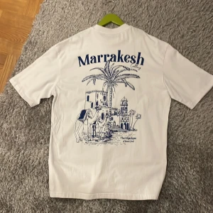 Vit Marrakesh t-shirt med tryck - Vit t-shirt med blått tryck på både bröstet och ryggen. På ryggen finns ett stort motiv med palmer, kameler och byggnader, samt texten 'Marrakesh'. T-shirten har rund hals och korta ärmar, perfekt för en chill och avslappnad stil.