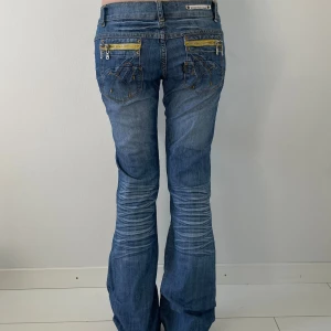 Blå bootcut jeans med dragkedjor - Snygga blå jeans med bootcut passform och låg midja. Jeansen har slitningar framtill, dekorativa sömmar och coola dragkedjor i gult på bakfickorna. Perfekta för dig som gillar detaljer och en avslappnad stil.