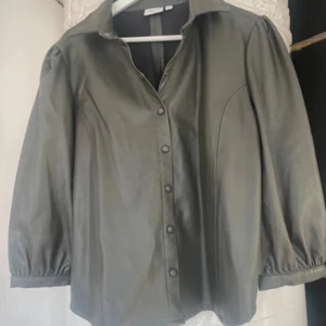 Grå skinnskjorta från Saint Tropez - Snygg grå skjorta i skinnimitation från Saint Tropez. Modellen har puffiga ärmar, klassisk krage och knappar framtill. Perfekt för dig som vill ha en edgy look med en touch av retro. Passformen är normal och materialet känns mjukt och följsamt.