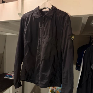 Svart overshirt från Stone Island - Svart overshirt från Stone Island med klassisk krage, dragkedja framtill och ikonisk patch på vänster ärm. Jackan har en bröstficka och är Snygg och stilren design som passar streetwear. Jackan kommer tyvärr inte längre till användning