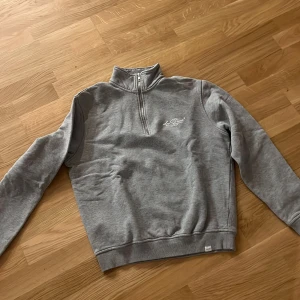 Grå half zip sweatshirt Les Deux - Grå sweatshirt från Les Deux med half zip och hög krage. Tröjan har lång ärm, broderad logga på bröstet och ribbade muddar. Perfekt för lager-på-lager och chill dagar.