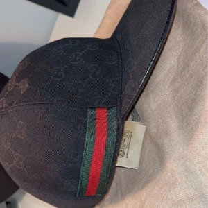 Svart keps från Gucci med GG-mönster - Snygg svart keps från Gucci med diskret GG-monogram över hela kepsen. Klassisk böjd skärm och ikoniska gröna och röda ränder på sidan. Tillverkad i bomullsmaterial och perfekt för dig som vill ha en stilren accessoar med lyxig känsla.tveka inte på att fråga 