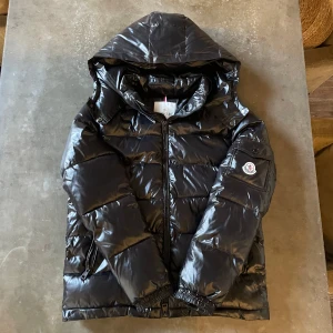 Moncler Maya - Snygg svart pufferjacka från Moncler med glansig finish och klassisk logga på ärmen. Jackan har huva, dragkedja framtill och två fickor med dragkedja. Perfekt för kalla dagar när du vill hålla stilen och värmen.