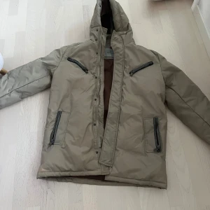 Beige Moncler dunjacka med huva - Snygg beige dunjacka från Moncler med huva och fyra dragkedjeförsedda fickor. Jackan har en clean look med diskret logga på ärmen och bruna detaljer på insidan. Jackan har lite defekter längst ner. Perfekt för kalla dagar när du vill hålla stilen och värmen.