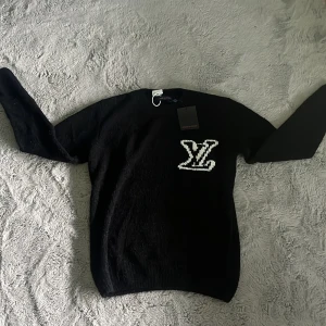 Svart stickad tröja Louis Vuitton - Svart stickad tröja från Louis Vuitton med stort vitt LV-logo på bröstet. Klassisk rund halsringning och långa ärmar. Perfekt för dig som vill ha en stilren men ändå lyxig look.