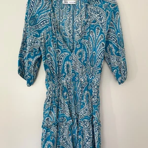 Blåvit mönstrad klänning från Zara - Supersnygg blå och vit kortklänning från Zara med paisleyinspirerat mönster. Klänningen har v-ringning, knyt i midjan och trekvartsärm. Tillverkad i ett lätt och luftigt material, perfekt för varmare dagar. Perfekt till sommaren, för stranden, middag,  till det mesta!! Använd fåtal gånger och är bra skick. 