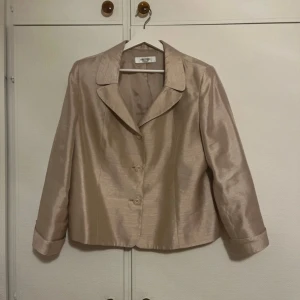 Champagnefärgad kavaj från Jones Studio - Snygg champagnefärgad kavaj från Jones Studio med glansig finish. Klassisk krage, tre knappar framtill och långa ärmar med uppvikta muddar. Perfekt för dig som vill sticka ut med en elegant och stilren look.