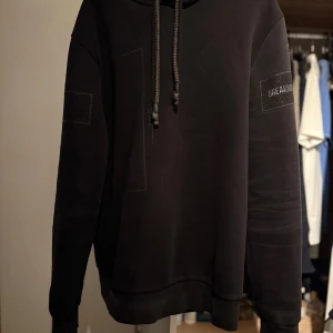 Svart hoodie med snörning Jack and jones  - Svart hoodie med snörning i huvan och diskreta patchdetaljer på ärmarna. Tröjan har en avslappnad passform och långa ärmar, perfekt för en cool och enkel stil.