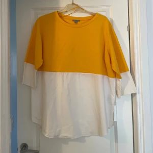 COS Gul och vit oversized tröja  - Säljer en oversized tröja från COS i storlek L. Tröjan har en cool färgblock-design med gul ovandel och vit nedandel, samt trekvartsärmar. Perfekt för dig som gillar en avslappnad och trendig stil.