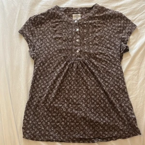 Brun babydoll topp  från L.O.G.G. - Säljer en brun gullig topp från L.O.G.G. med vitt blommigt mönster. Modellen har korta ärmar, rund halsringning och knappar framtill. Tillverkad i mjuk bomull.