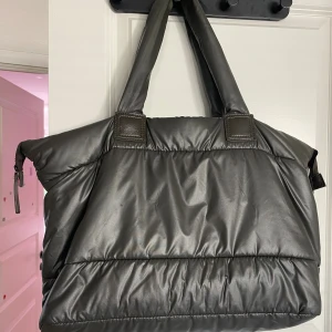 Stor svart axelväska Duke & Duchess - Säljer en stor och mjuk axelväska från Duke & Duchess Marshmallow Bag Collection. Väskan är svart med quiltad look, har breda handtag och dragkedja upptill. Insidan är brun med innerfack och en snygg metallbricka med logga. Perfekt för dig som gillar oversized och praktiskt.