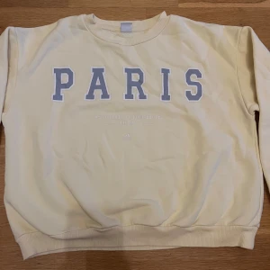 Ljusgul/smör sweatshirt med PARIS från Lindex - Ljusgul tröja från Lindex i storlek 146/152. Framsidan har ett stort tryck med texten PARIS i grått och vitt. Tröjan har rund hals, ribbade muddar och är tillverkad i mjuk bomull.