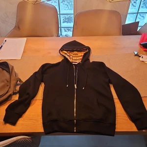 Svart Burberry hoodie med dragkedja - Svart hoodie från Burberry med klassiskt rutigt foder i huvan. Tröjan har dragkedja framtill, snörning vid huvan och är tillverkad i 100% bomull. Perfekt för dig som gillar stilrena och exklusiva plagg med snygga detaljer.