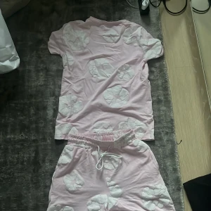 Rosa denim tears set - Säljer ett par ljusrosa shorts med vita blommönster och elastisk midja med snörning. De är tillverkade i mjuk bomull och har en avslappnad passform, perfekta för varma dagar eller chilla hemma.