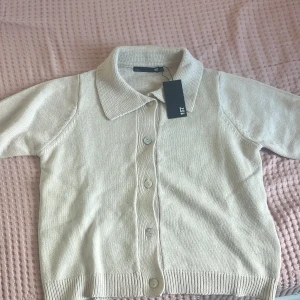 Beige kofta från 157 med krage - Snygg beige kofta från 157 med klassisk krage och knäppning framtill. Koftan har korta ärmar och ribbade muddar vid nederkant och ärmslut. Perfekt för lager-på-lager och enkel att matcha till olika outfits.