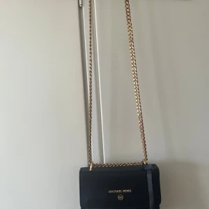 Svart axelväska från Michael Kors - Snygg svart axelväska från Michael Kors med guldfärgad kedjerem och logga framtill. Väskan har ett stilrent och klassiskt utseende, magnetlås och innerfack med dragkedja. Perfekt storlek för det viktigaste och passar till många outfits.