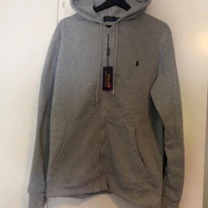 Grå hoodie från Polo Ralph Lauren - Snygg grå hoodie från Polo Ralph Lauren med dragkedja, huva och snörning. Klassisk broderad logga på bröstet och två stora fickor framtill. Perfekt för en chill och stilren look.