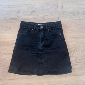 Svart jeanskjol med fickor - Snygg svart jeanskjol med klassisk femficksdesign och knappstängning fram. Kjolen har en lätt A-linjeform och är kort i modellen. Perfekt att matcha med sneakers eller boots för en cool look.