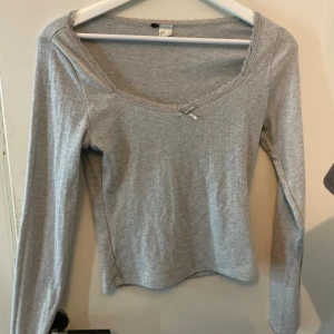 Grå ribbad långärmad topp med spets från H&M - Säljer en ljusgrå ribbad topp med lång ärm och söt spetskant runt den breda ringningen. Liten rosett framtill ger en gullig touch. Toppen är i mjuk bomullsmix och har en slim passform som sitter snyggt på kroppen. Andvänd ett par fåtals gånger! 