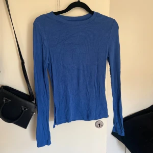 Blå ribbad långärmad tröja - En basic blå långärmad tröja i ribbad bomull med slitsar i sidorna. Tröjan har en rund halsringning och sitter ganska tight mot kroppen. Perfekt att styla med jeans eller kjol för en enkel och clean look.