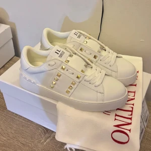 Valentino Rockstud vita sneakers(Nya) - Snygga vita Valentino Rockstud sneakers i skinn med ikoniska guldiga nitar längs sidorna och på hälen. Klassisk rund tå, platt sula och breda vita skosnören. Perfekta för dig som vill ha en clean look med lite edge.