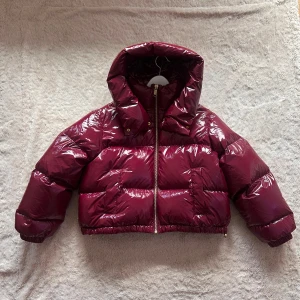Vinröd pufferjacka från Michael Kors - Säljer en glansig vinröd pufferjacka från Michael Kors med huva och guldfärgad dragkedja. Jackan har MK-logga på ärmen och ett stort broderat MK-märke på ryggen. Croppad modell med elastiska muddar och quiltad design.  Skick: Nyskick då den endast är testad på. Stolek: (M)💜