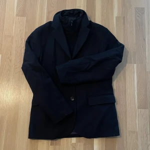 Marinblå jackkavaj från mango - Stilren marinblå kavaj med klassisk krage och två knappar framtill. Kavajen har två fickor med lock och en innerkrage med dragkedja för extra detalj. Perfekt för dig som gillar en enkel och snygg look.