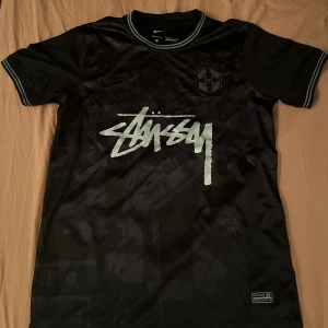 Svart Brasil x Stüssy x Nike fotbollströja - Unik svart fotbollströja från Nike i samarbete med Stüssy och Brasil. Tröjan har korta ärmar, Dri-FIT-material och stora Stüssy-loggan framtill. Broderat Brasil-märke på bröstet och randiga muddar vid hals och ärmslut. Perfekt för dig som gillar streetwear och fotboll.