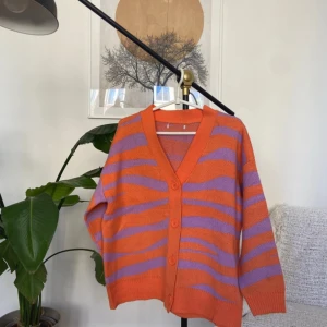 Orange och lila kofta med zebramönster - Färgstark kofta med orange och lila zebramönster. Koftan har v-ringning, knappar framtill och är stickad i ett mjukt material. Passformen är loose och ärmarna är långa, perfekt för dig som vill sticka ut med en cool och trendig look.