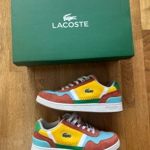 Lacoste x Peanuts sneakers strl 39 - Unika Lacoste x Peanuts T-Clip sneakers i färgglad mix av gult, grönt, blått, rosa och vitt. Skorna har mockadetaljer, perforerad tå, snörning och klassisk krokodillogo på sidan. Använda en handfull gånger och levereras i orginalkartongen. Köpt för 1300 i Lacoste butiken i Täby.