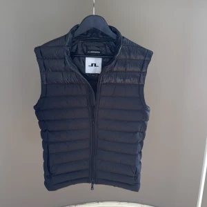 Svart dunväst från J.Lindeberg - Snygg svart dunväst från J.Lindeberg med quiltad design och dragkedja framtill. Västen har en stilren look med hög krage och är perfekt att slänga över en hoodie eller tröja. Passar dig som gillar sportig och clean stil. (Använd max 10 gånger)