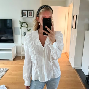 Vit blus med volangkrage och spets - Supersöt vit blus med stor volangkrage och broderade spetsdetaljer. Långa ärmar med manschett och knappar framtill. Perfekt för dig som gillar romantisk och trendig stil. Luftig passform och snygg till både jeans och kjol.