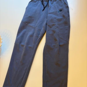 Blå mjukisbyxor från Nike - Snygga blå mjukisbyxor från Nike med svart logga på vänster lår. Byxorna har dragsko i midjan och praktiska fickor, varav en med dragkedja. Perfekta för chill eller träning, tillverkade i mjukt bomullsmaterial.